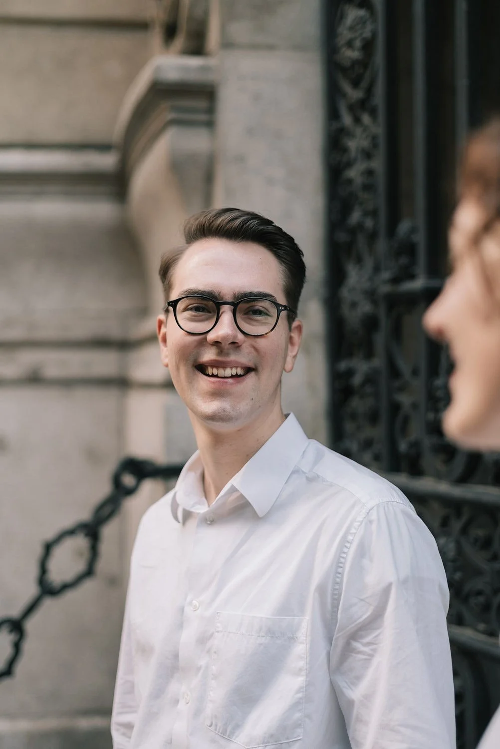 Homem sorrindo com óculos de grau, vestindo camisa branca, ao lado de uma pessoa parcialmente visível, em ambiente urbano.