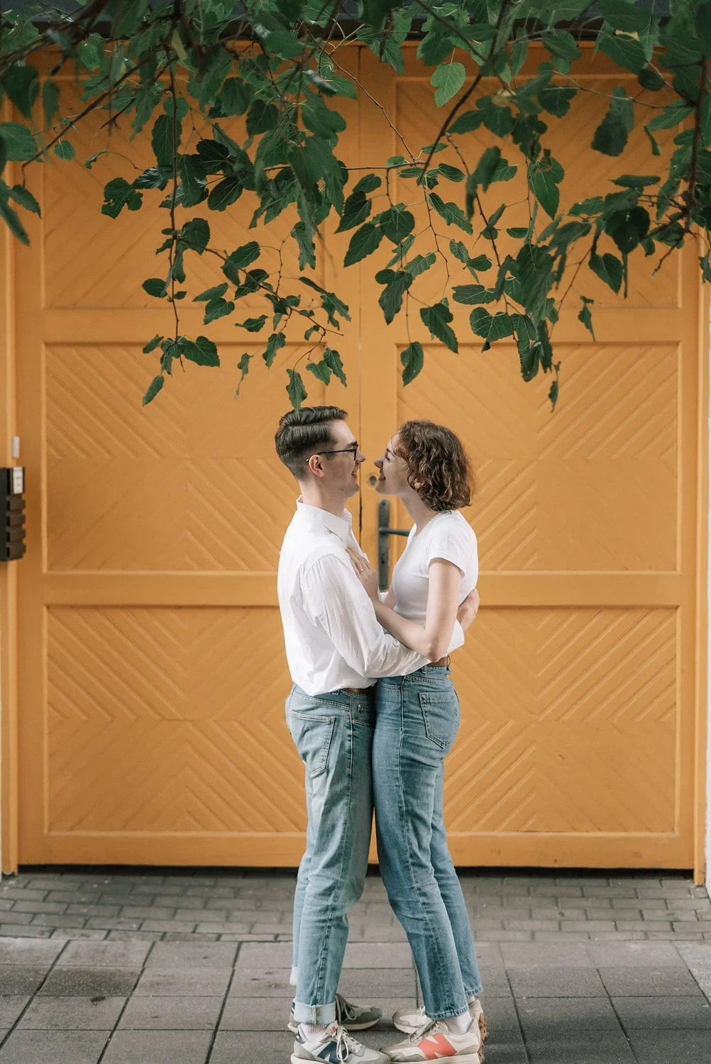 Um casal jovem, de camiseta branca e jeans, se abraçando na frente de uma porta de madeira, com folhas verdes penduradas acima deles.