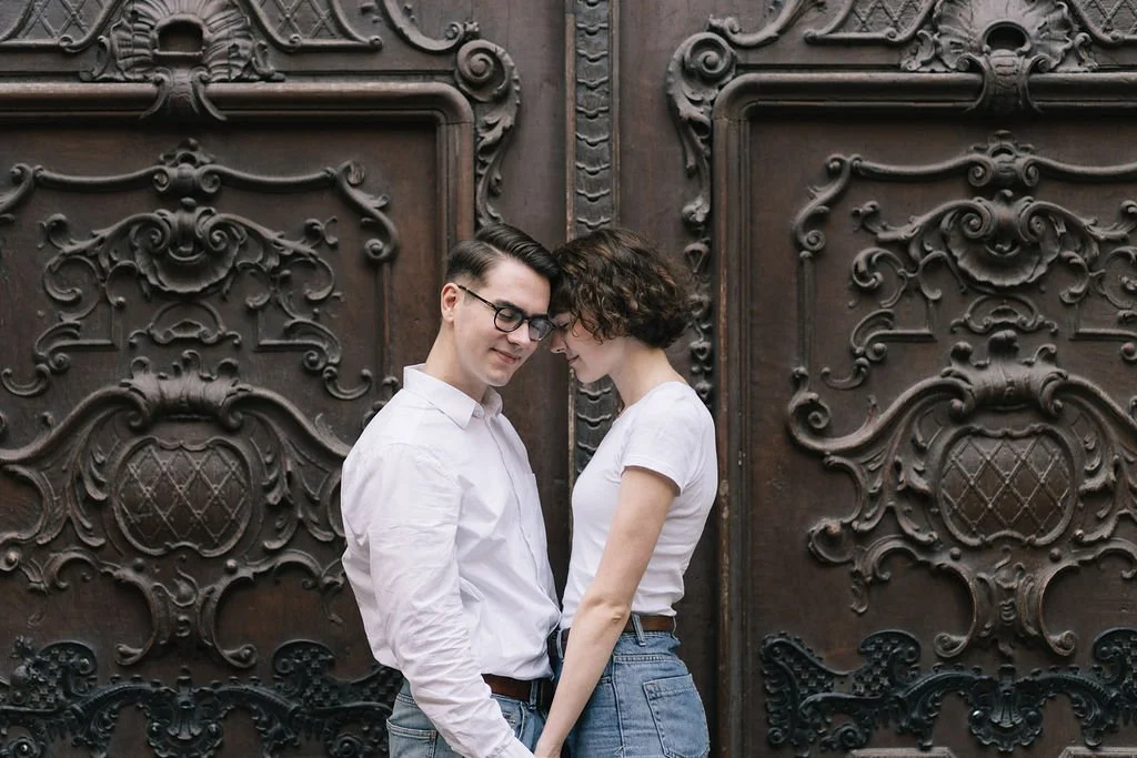 Um casal jovem está de cabeça juntada, com expressão tranquila, em frente a uma porta de ferro com detalhes ornamentais escuros.