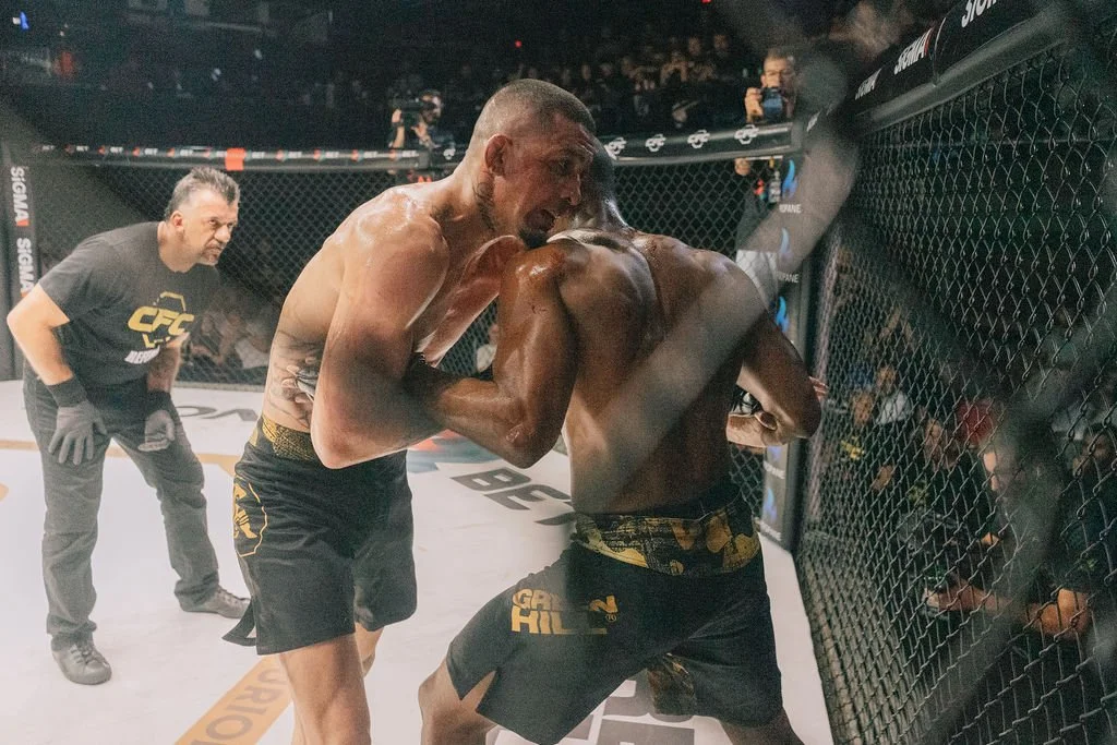 Dois lutadores de MMA em combate próximo na jaula, um de rosto visível, o outro com costas viradas, enquanto um árbitro observa atentamente ao lado.