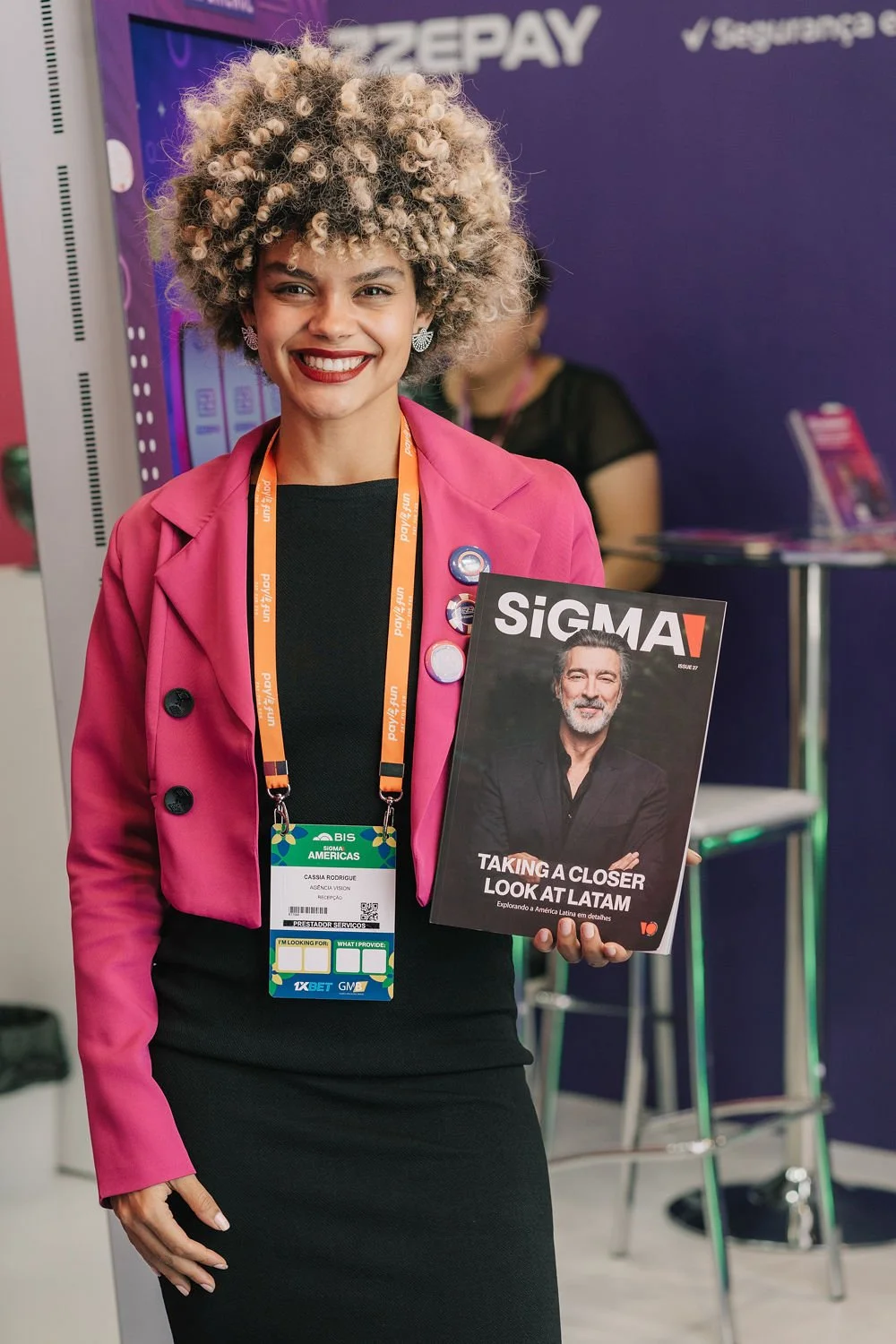 Mulher sorridente usando blazer rosa e camiseta preta, segurando uma revista intitulada 'SIGMA' com a capa mostrando um homem de barba cinza.