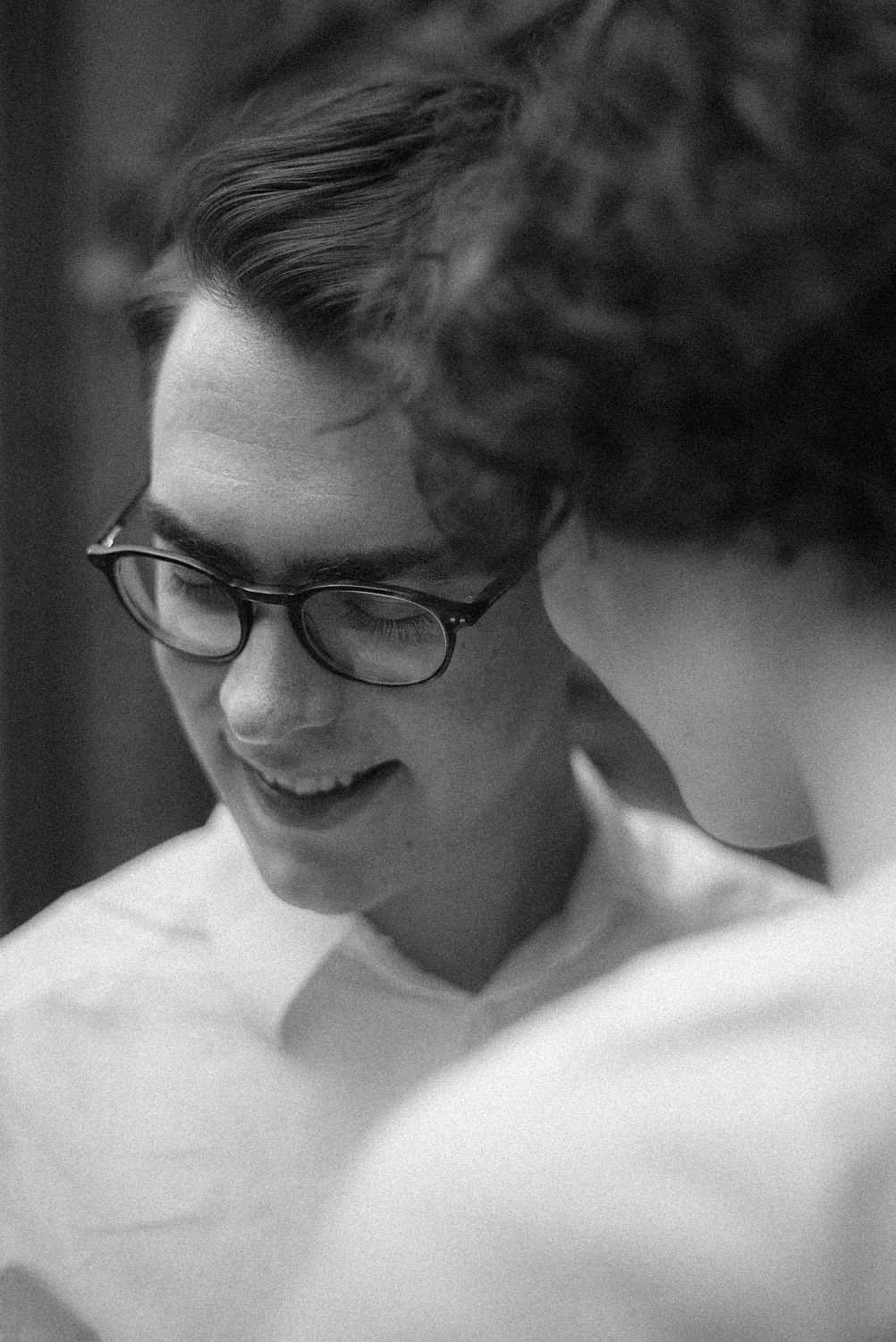 Duas pessoas próximas, uma com óculos, sorrindo e olhando para o lado, em um ambiente interno. Preto e branco.