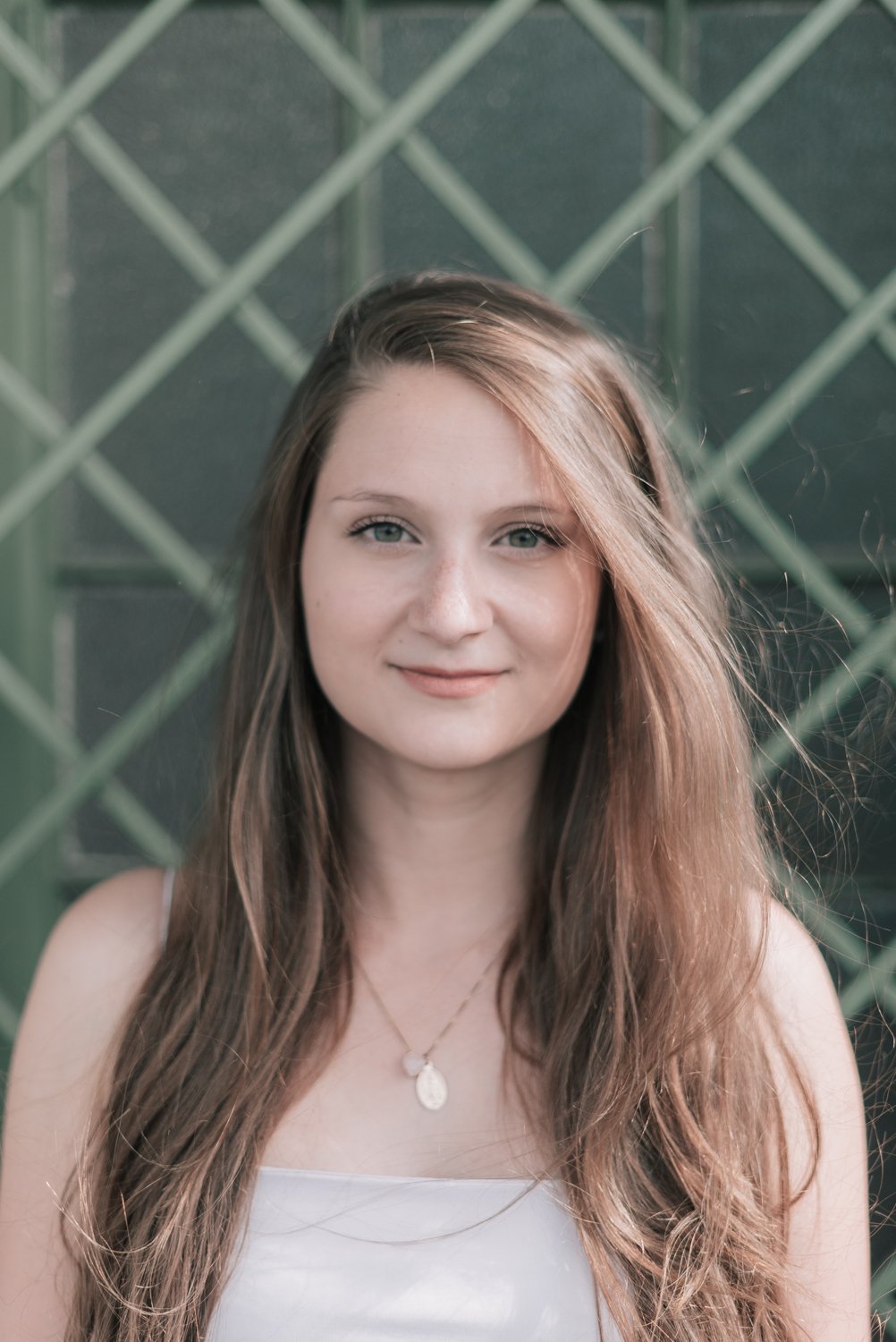 Jovem mulher com cabelo longo castanho claro, usando vestido branco, com colar, sorrindo na frente de uma cerca de ferro verde.