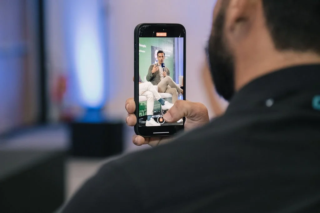 Pessoa gravando video de uma mulher falando em um evento ou palestra, vista de costas, segurando um celular.