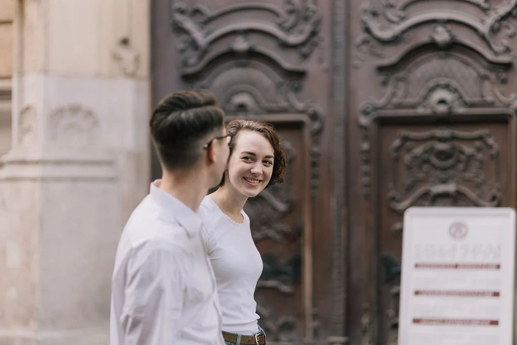 Um homem e uma mulher sorrindo e conversando na rua, na frente de uma porta de madeira ornamentada.