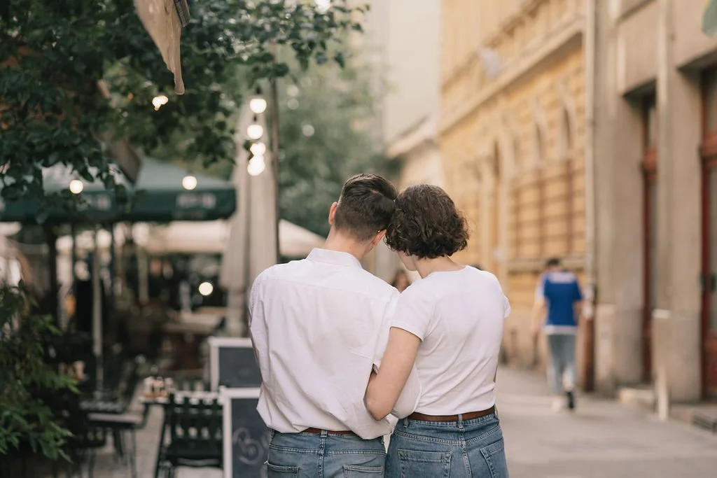 Um casal jovem de costas, usando roupas brancas e jeans, abraçado na rua de uma cidade, com prédios antigos ao fundo e pessoas ao longe.