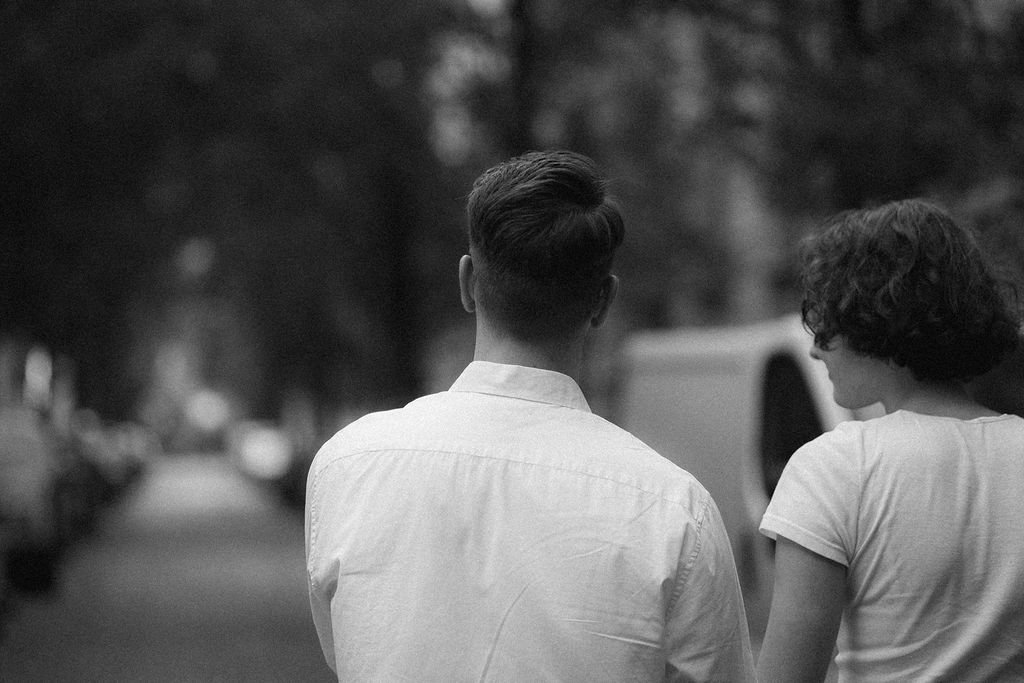 Homem e mulher andando de costas por uma rua em um ambiente urbano com árvores ao redor, em uma fotografia em preto e branco.