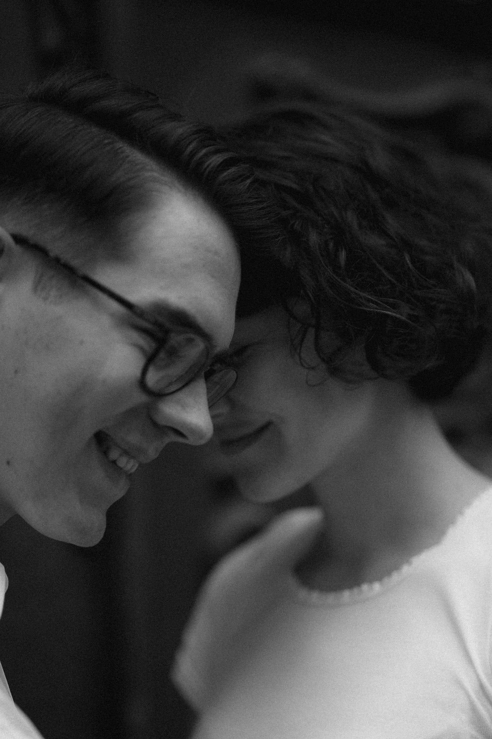 Imagem em preto e branco de duas pessoas, uma com óculos e cabelo liso, e outra com cabelo cacheado, com as suas testas tocando e sorrindo.