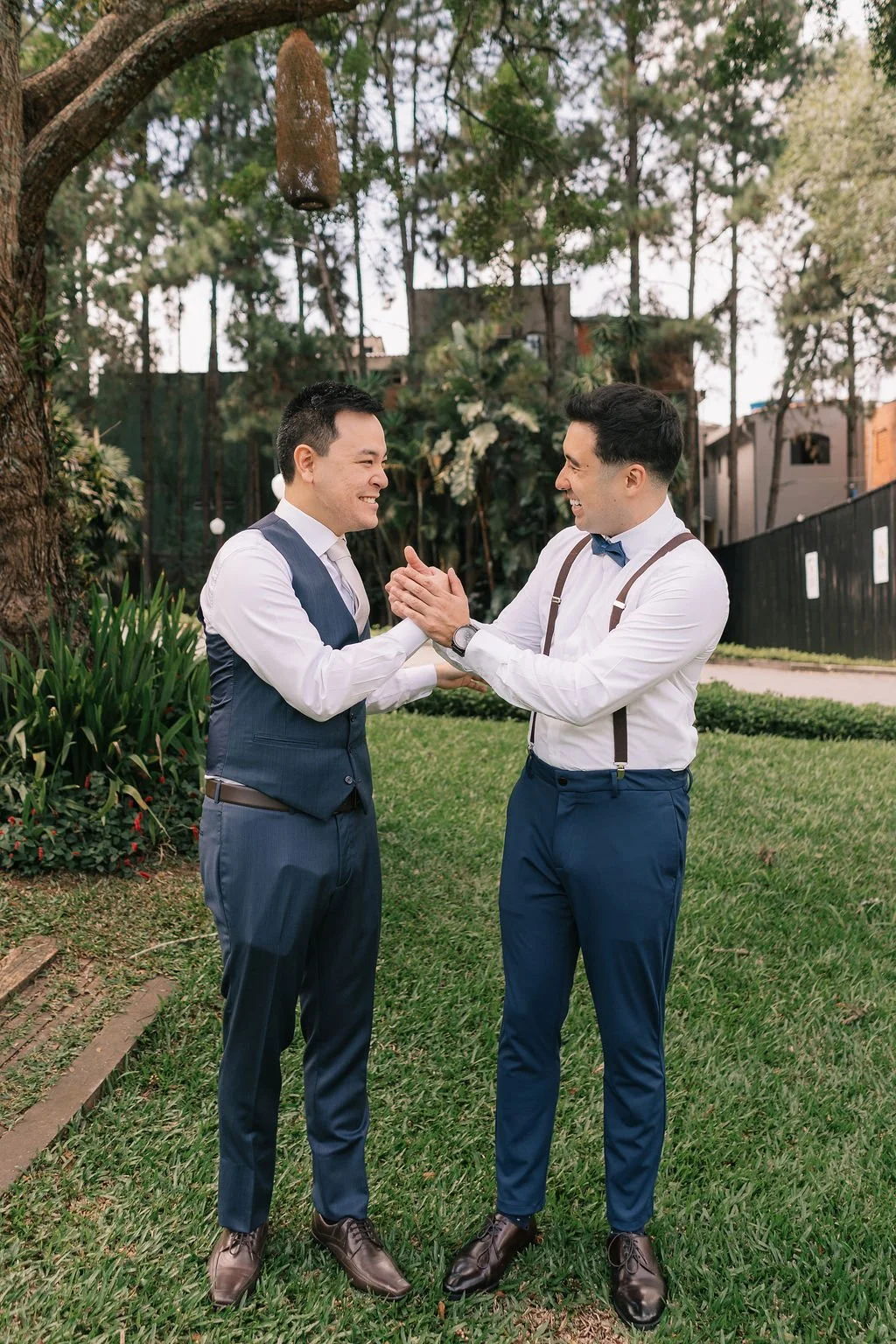 Dois homens sorridentes vestindo roupas sociais brancas e calças azul escuro, moldando-se com as mãos em meio a um gramado verde sob uma árvore, em ambiente externo