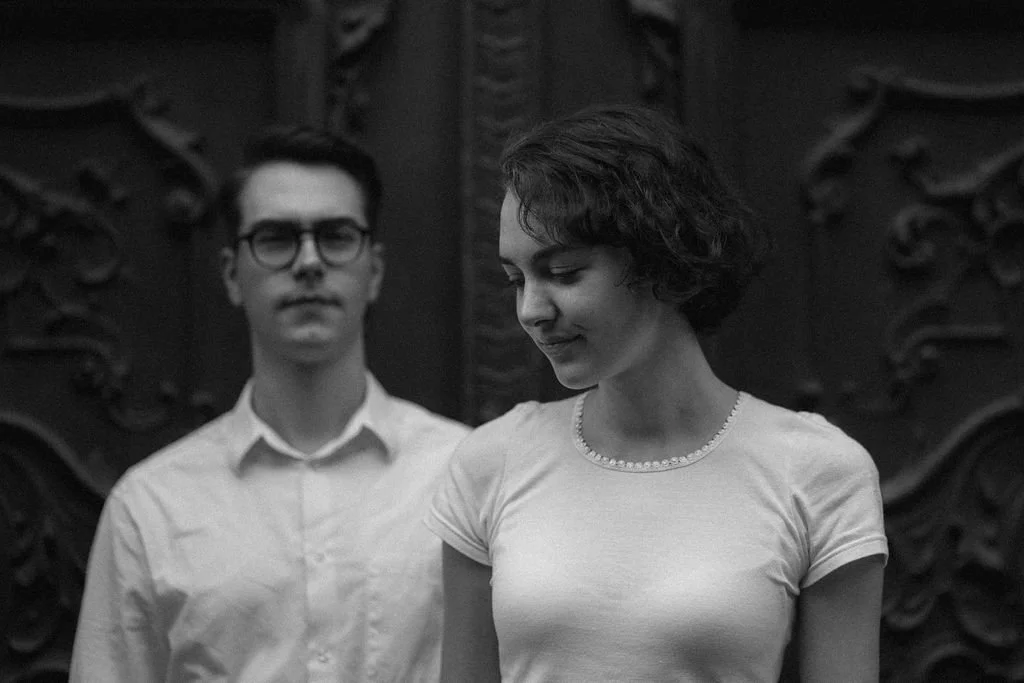 Homem e mulher em um ambiente de fundo decorado com detalhes em relevo, ambos com expressão neutra, mulher ao centro olhando para baixo.