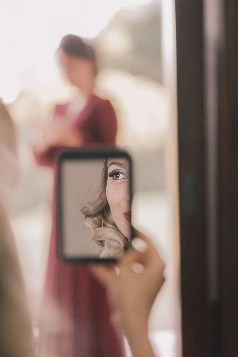 Mãe tirando uma selfie no espelho com a filha ao fundo, que aparece refletida no espelho, com cabelo cacheado e maquiagem vermelha.