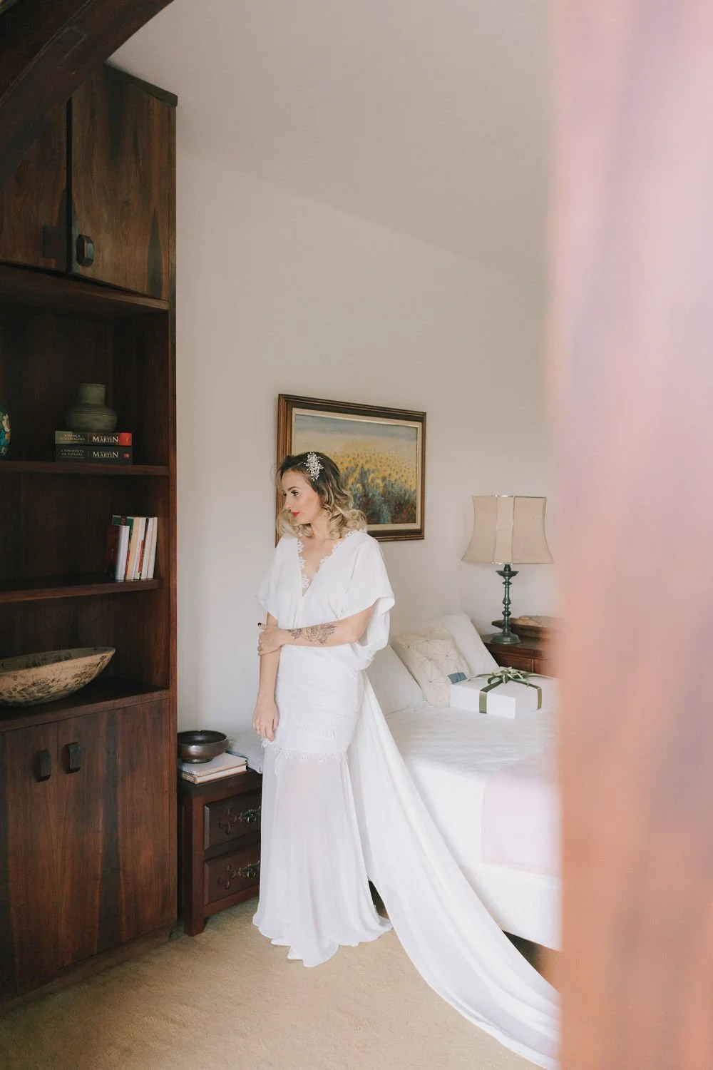 Mulher em vestido branco em um quarto, segurando o braço, ao lado da cama com uma embalagem de presente