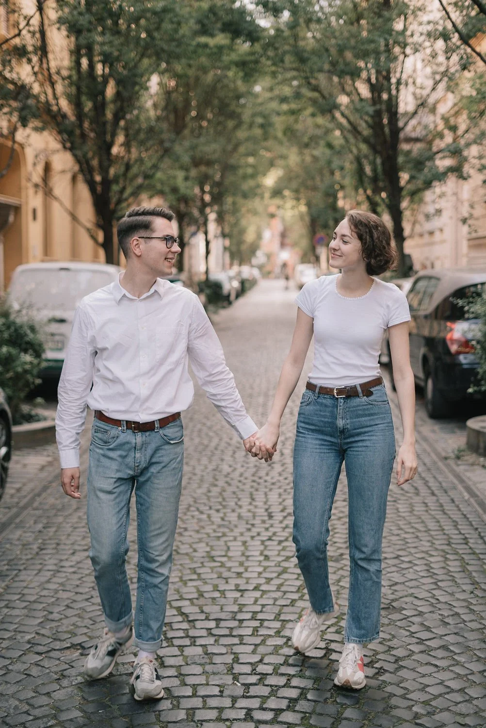 Casal jovem caminhando de mãos dadas na rua com árvores e carros estacionados ao lado, durante o pôr do sol.