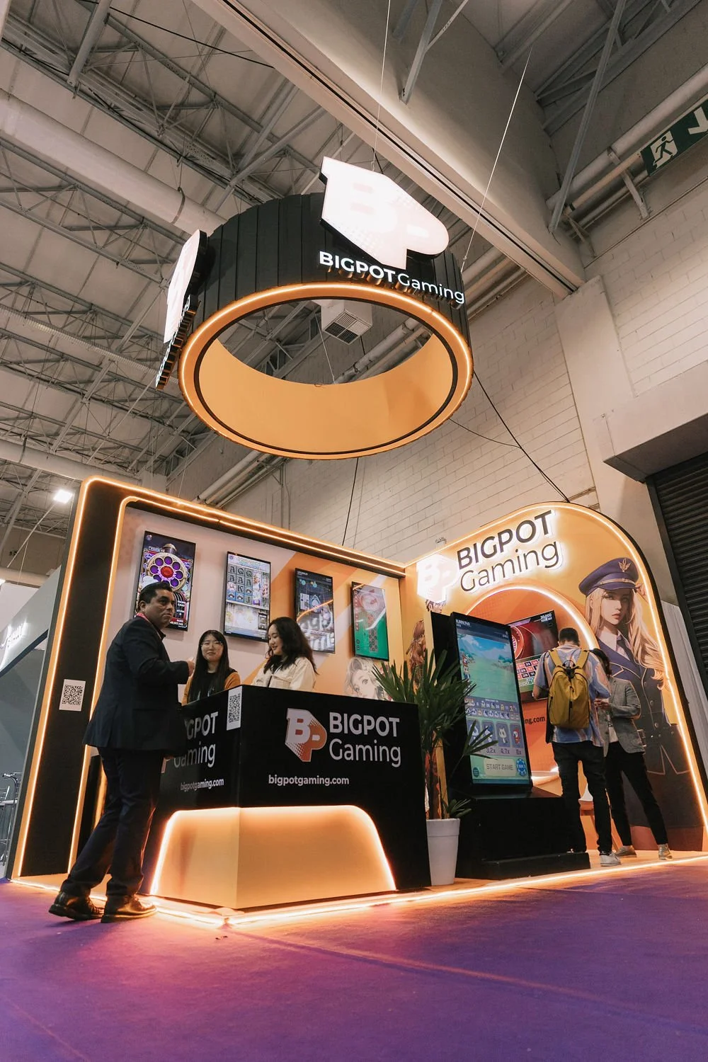 Estande da BIGTOP Gaming em uma feira, com pessoas conversando e jogando. Decoração moderna com luzes de néon, telas com jogos e uma estrutura de tronco suspensa no teto com o logotipo da loja.