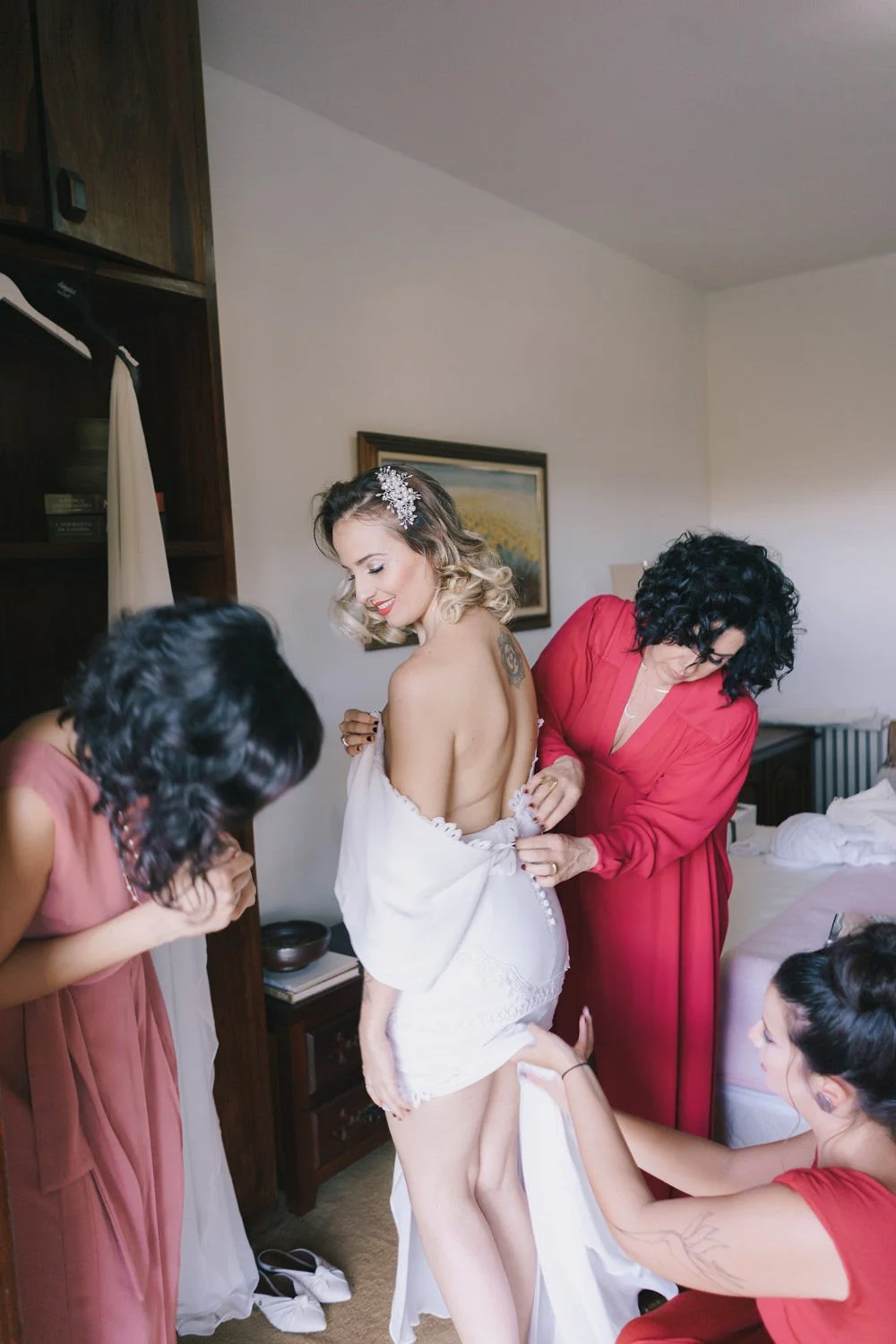 Mulher de vestido branco sendo ajudada por amigas a vestir seu vestido de casamento, em um quarto.