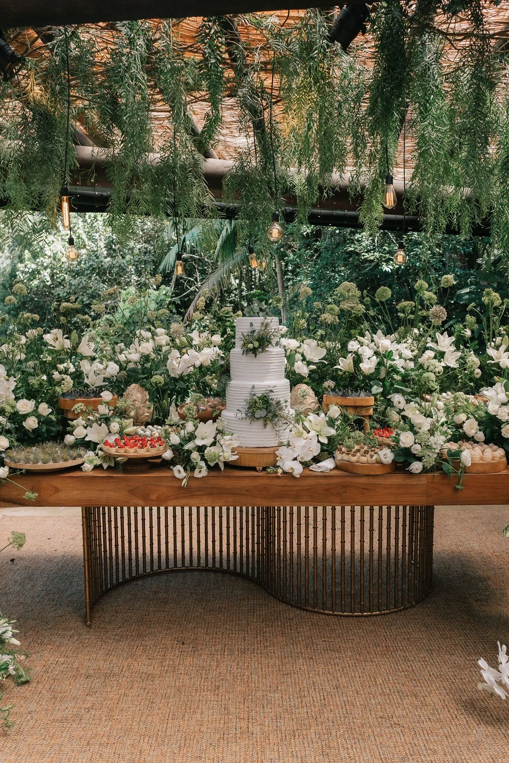 Mesa de festa de casamento decorada com flores brancas, bolo de vários andares com detalhes verdes e luzes penduradas, em ambiente com muitas plantas e vegetação ao redor.