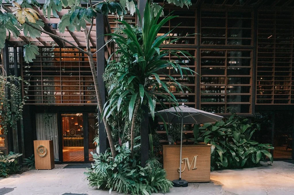 entrada de restaurante ou café com fachada de vidro, plantas tropicais e uma mesa com guarda-sol