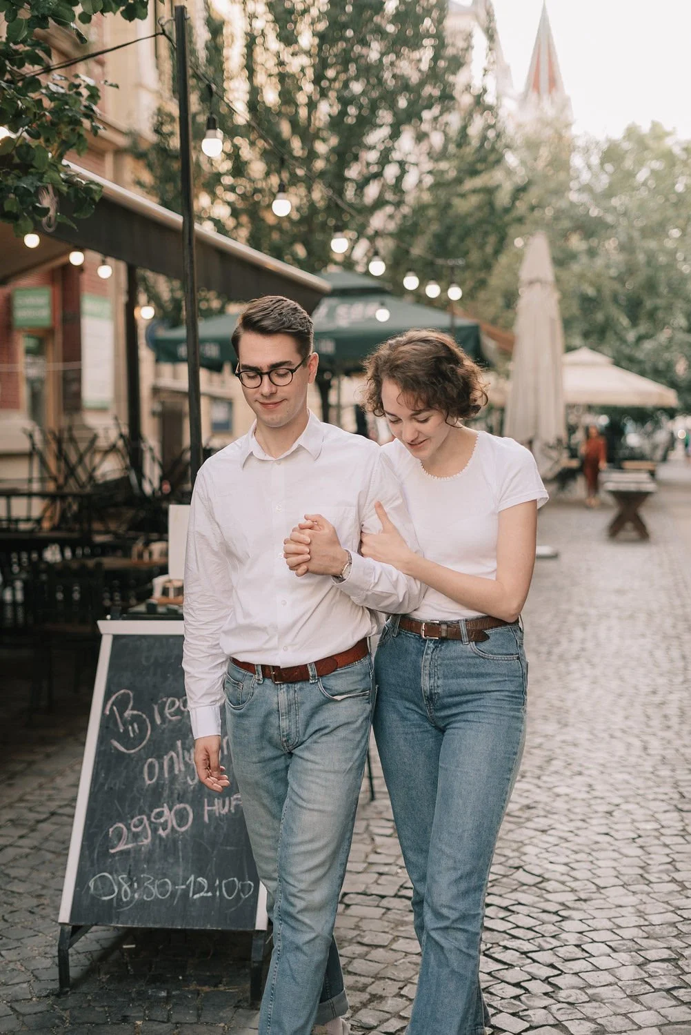 Casal caminhando de mãos dadas na rua ao pôr do sol, com decoração de luzes e um quadro de aviso ao fundo.