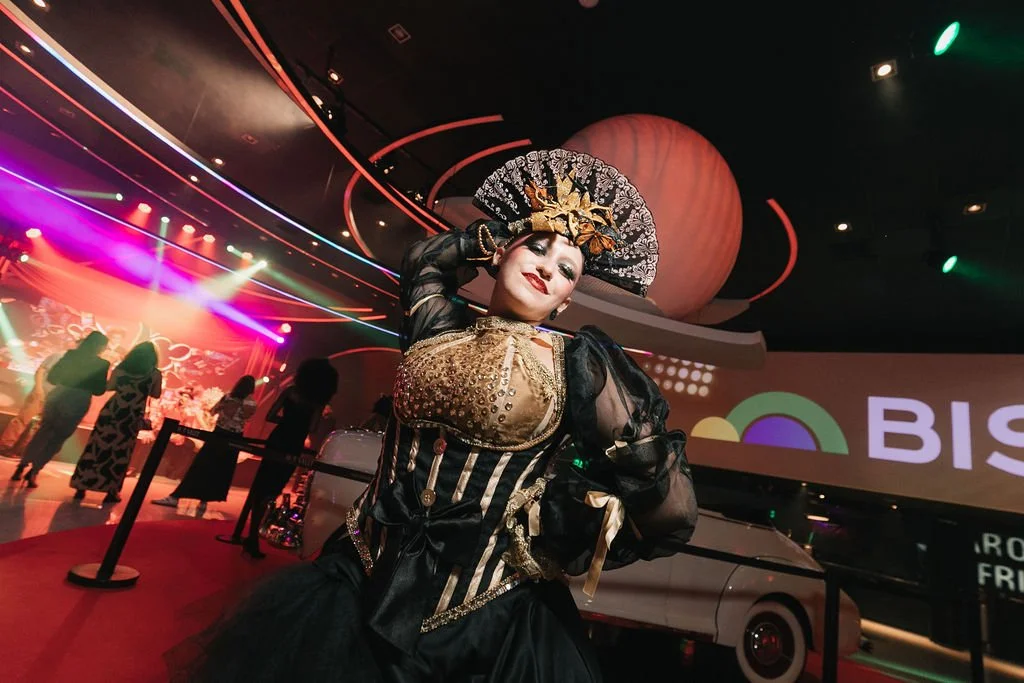 Pessoa com fantasia de balé, maquiagem elaborada, usando um vestido preto e dourado com detalhes de saia rodada, em um evento com iluminação de nightclub, com um fundo de palco, uma grande tela com a palavra 'BIS' e várias pessoas ao fundo.