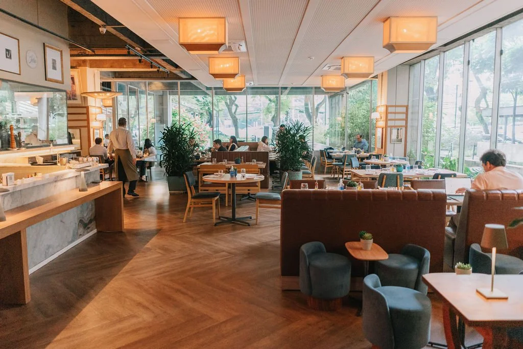Interior de restaurante com mesas, cadeiras e iluminação suave, com clientes e funcionários, e janelas que deixam a luz natural entrar.