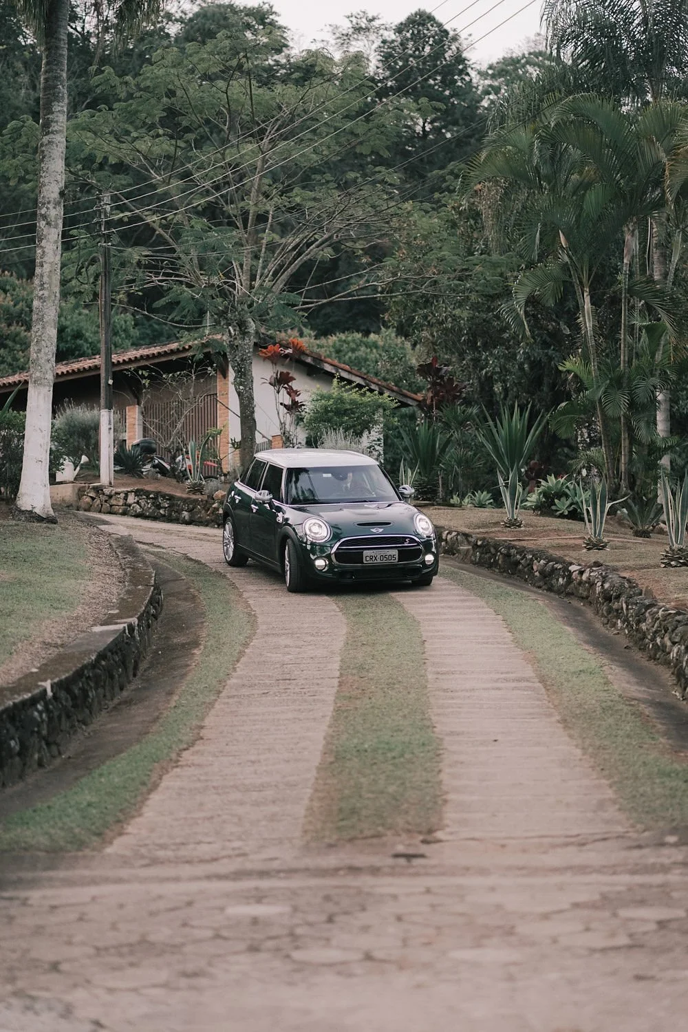 Carro preto estacionado em uma estrada de terra rodeada de plantas e árvores em uma área rural, com uma casa ao fundo.