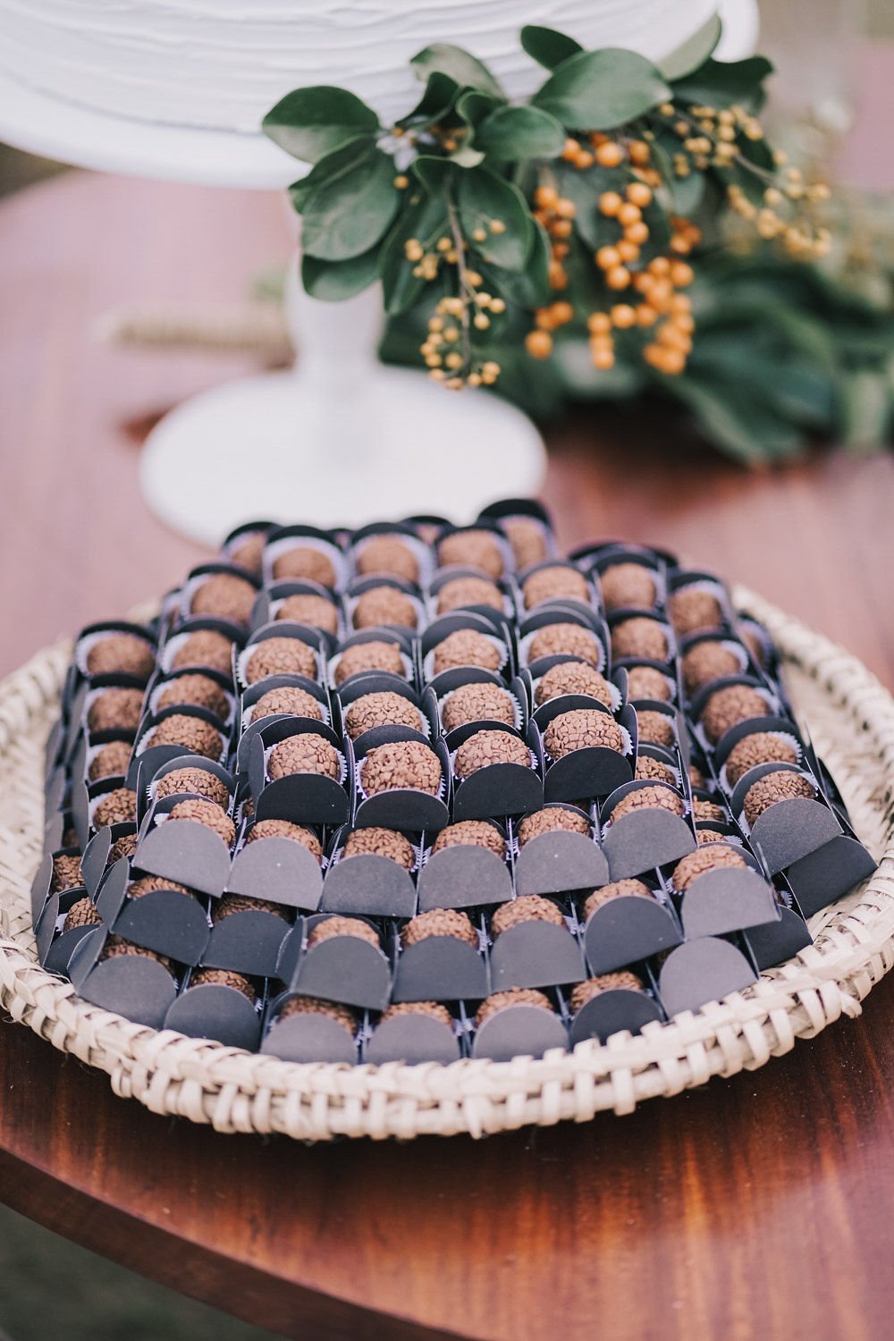 Trilho de docinhos de brigadeiro em uma bandeja de palha, com decoração de folhas verdes e pequenos frutos amarelos ao fundo