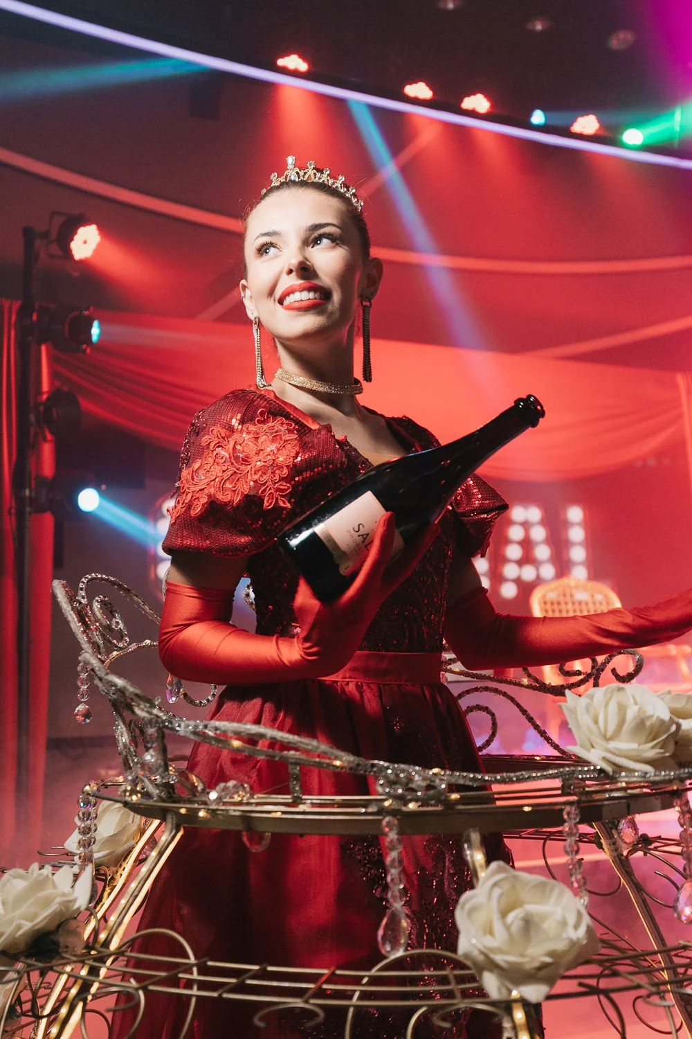 Mulher vestida com vestido vermelho, usando uma tiara e brincos, segurando uma garrafa de espumante em um ambiente com luzes coloridas e decoração festiva.