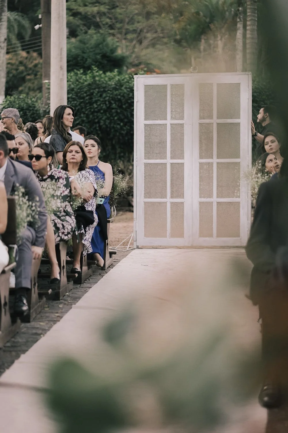 Pessoas assistindo a cerimônia de casamento ao ar livre, com uma porta de madeira branca ao fundo, em um ambiente natural.
