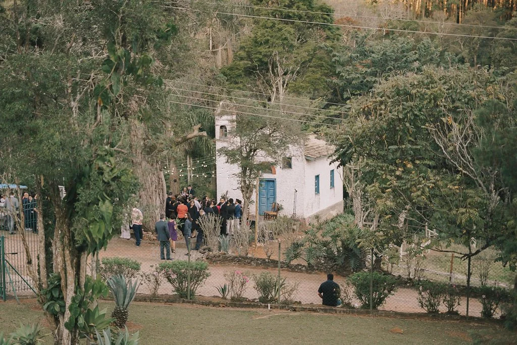 Reunião ao ar livre em frente a uma capela branca rodeada por árvores, com várias pessoas conversando e uma pessoa sentada na cerca.