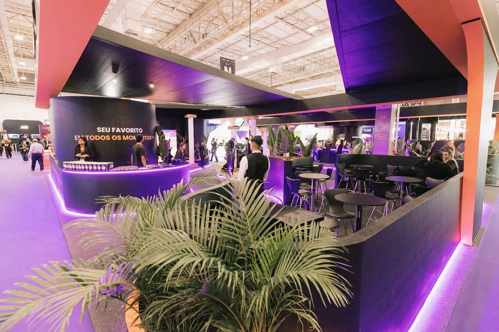 Bar com iluminação roxa e cadeiras pretas, com pessoas sentadas e um atendente atrás do balcão, em um espaço interno com plantas decorativas.