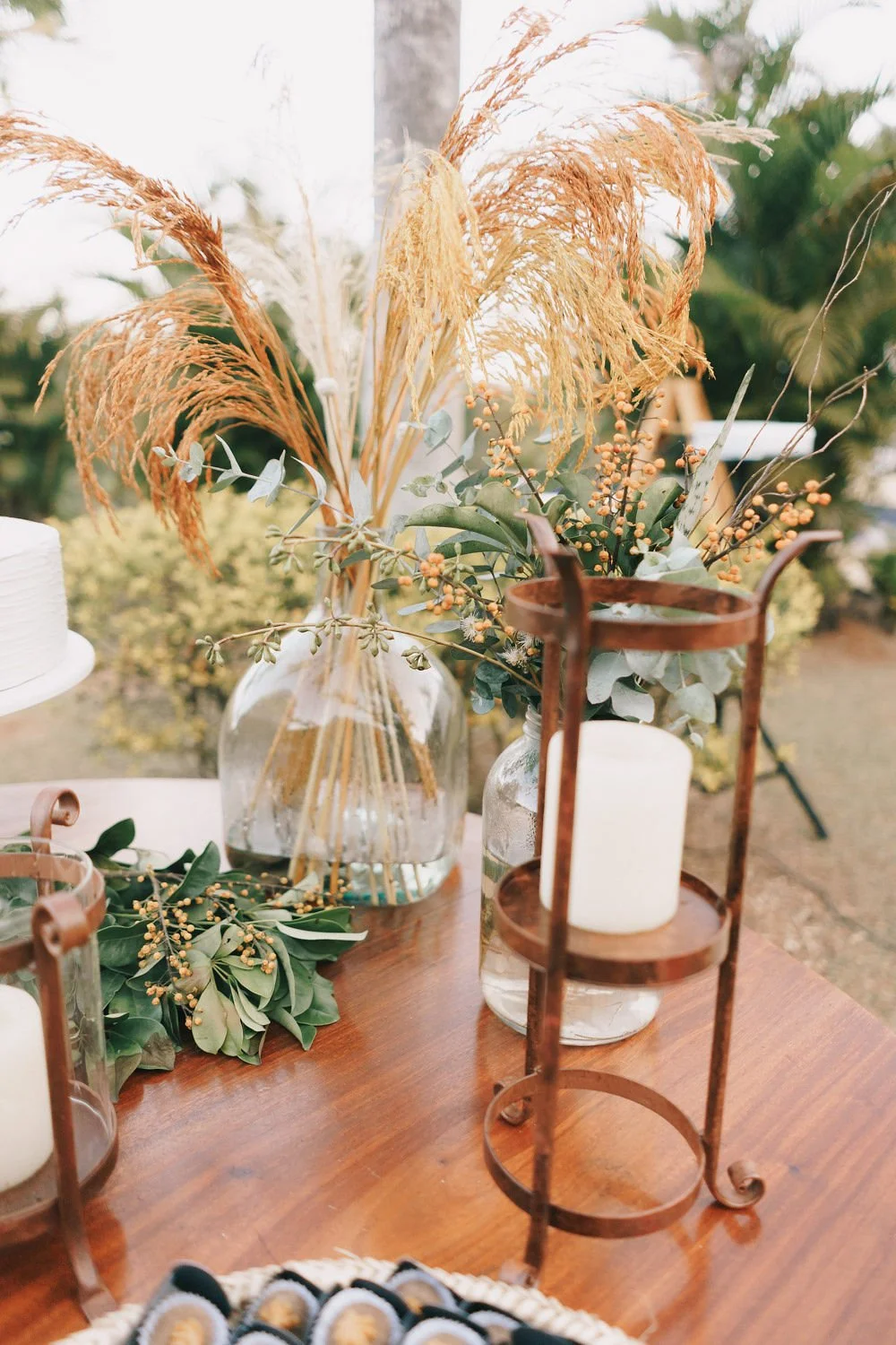 Arranjo decorativo com folhas verdes, galhos e plumas secas em vasos de vidro, sobre uma mesa de madeira, com velas brancas em suportes de ferro ao redor, em ambiente ao ar livre.