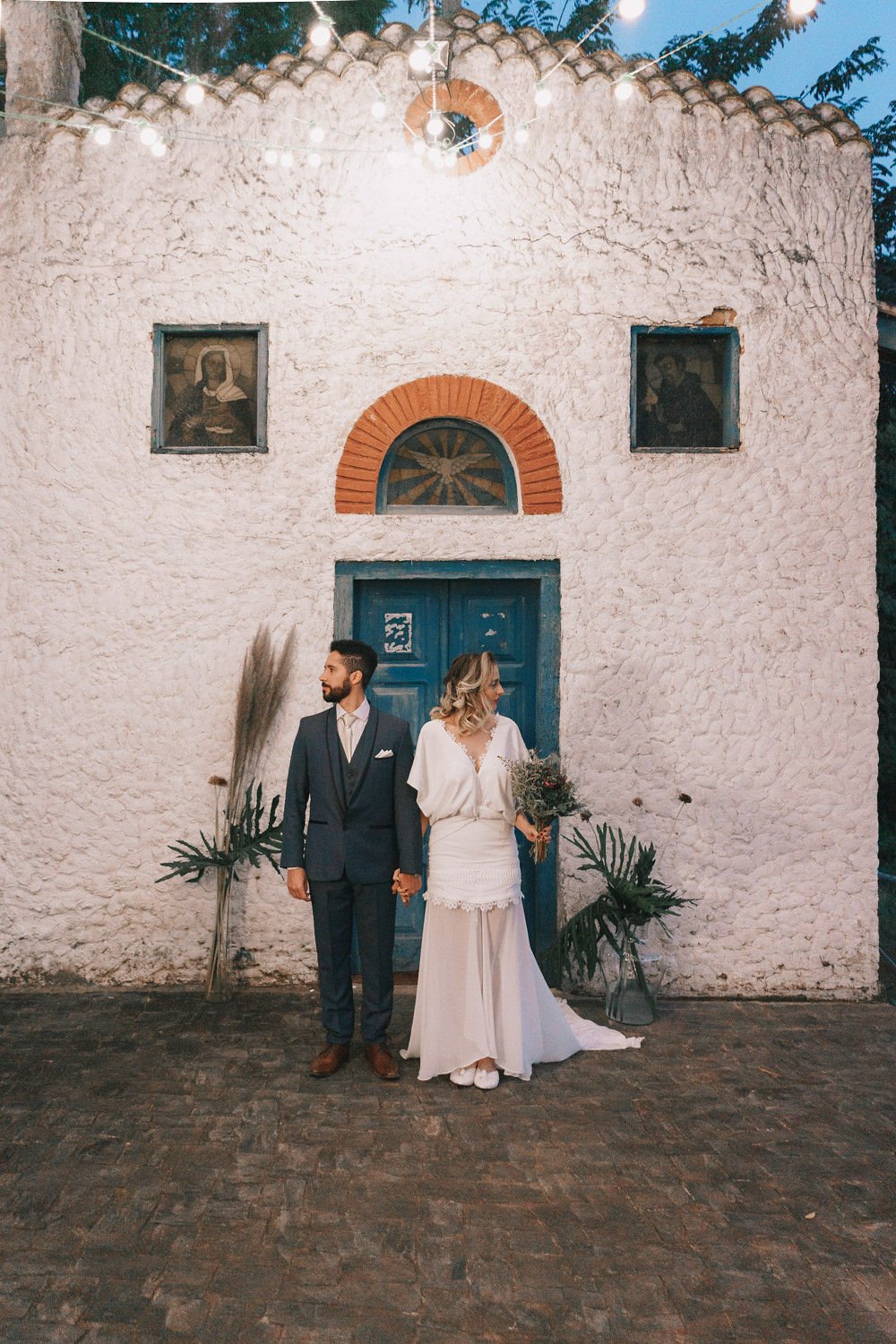 Casal de casamento segurando as mãos em frente a uma igreja de paredes brancas e porta azul, decorada com plantas e iluminação ambiente.
