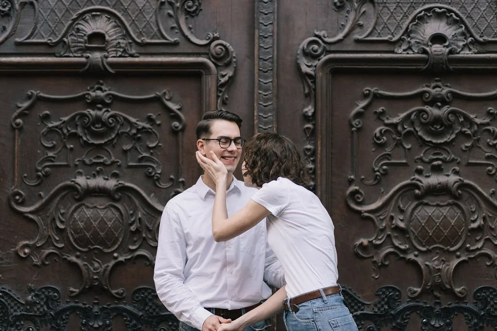 Um casal sorridente brincando na rua, com uma porta de ferro ornamental ao fundo.