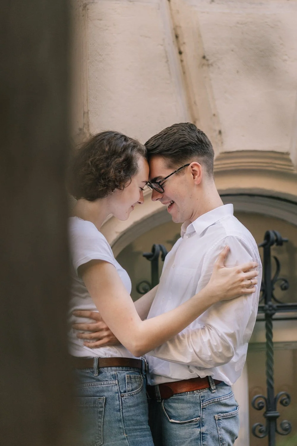 Casal com testa juntos sorrindo, segurando um ao outro em um ambiente externo com parede de pedra ao fundo.