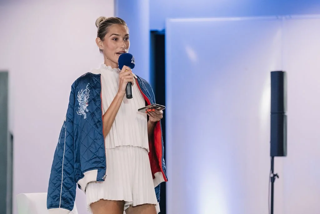 Mulher jovem falando ao microfone em um evento, segurando um celular, com fundo de parede branca e equipamento de som ao lado.