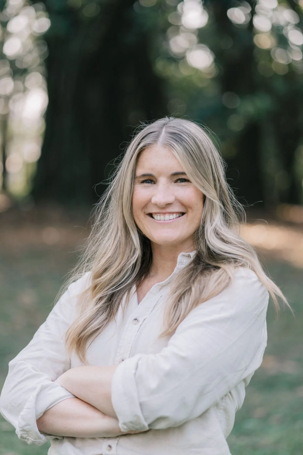 Mulher sorridente com cabelo louro claro, vestindo blusa branca, em ambiente ao ar livre com árvores ao fundo.