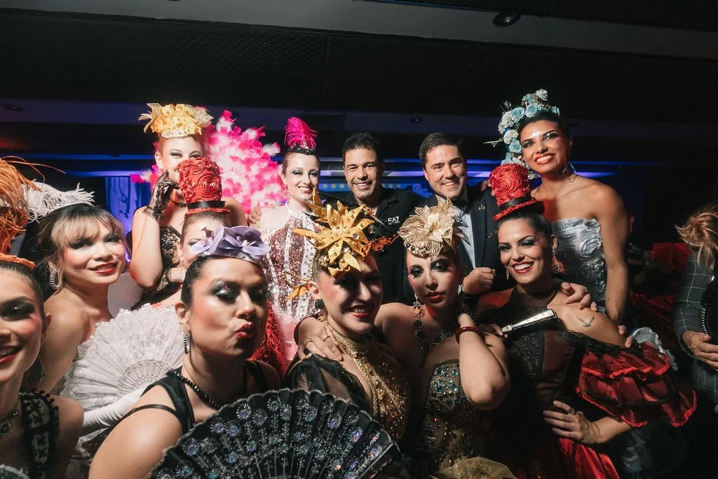 Grupo de pessoas vestidas com roupas de fantasia, muitas com acessórios elaborados e cabelo estilizado, em um evento com iluminação colorida.