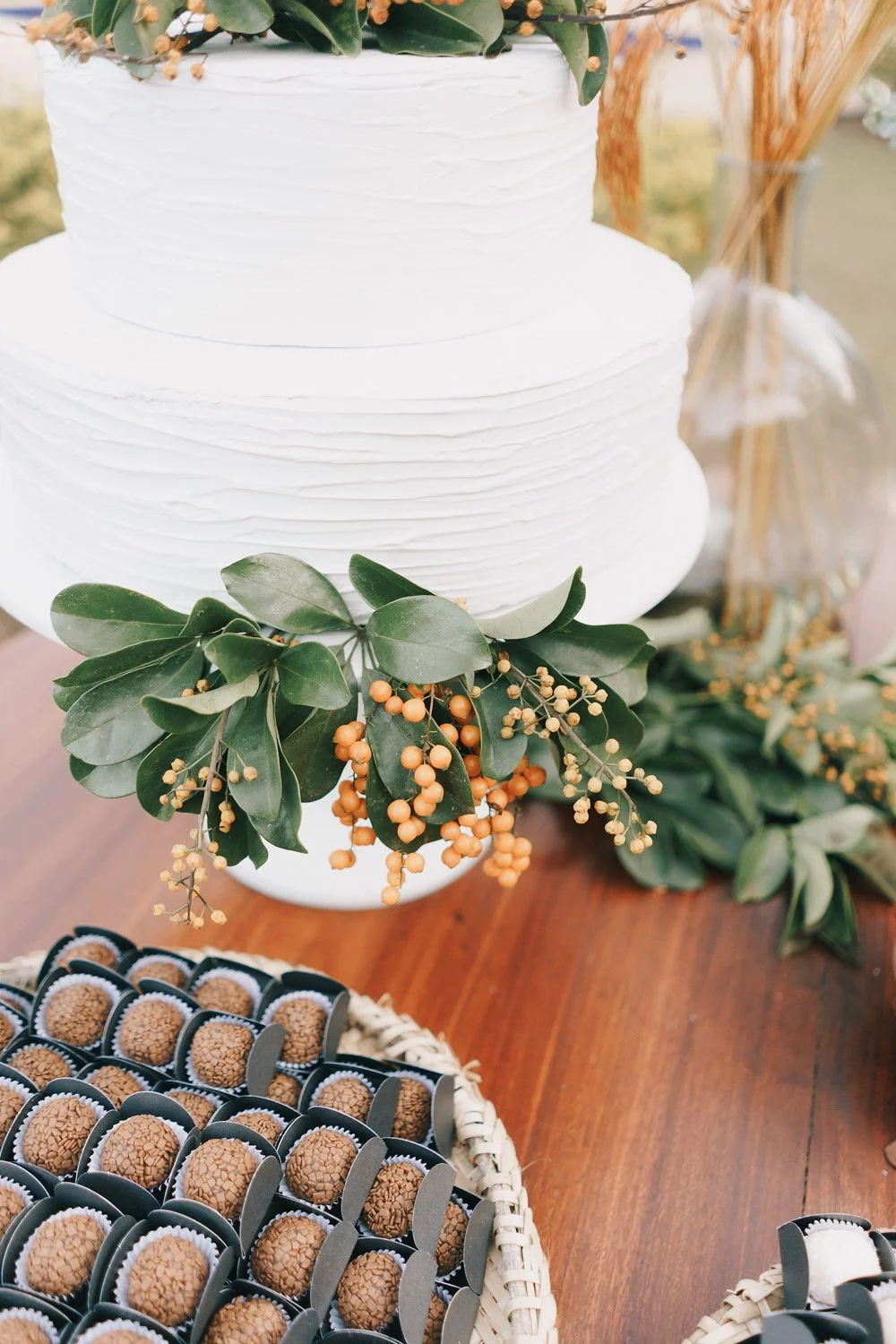 Bolo de casamento branco decorado com folhas verdes e flores laranjas, acompanhado de doces de chocolate ao leite em uma bandeja, em uma mesa de madeira.