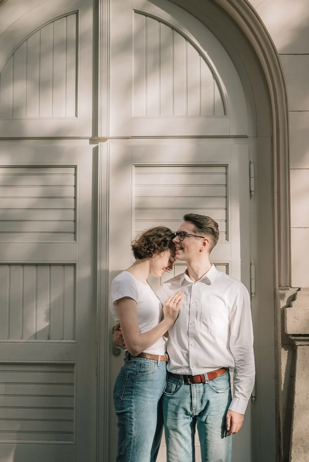 Casal sorrindo, com a mulher encostada no homem, em frente a uma porta branca de madeira. Ela usa camiseta branca e jeans, ele usa camisa branca e jeans. Estão se aproximando com expressão afetuosa.