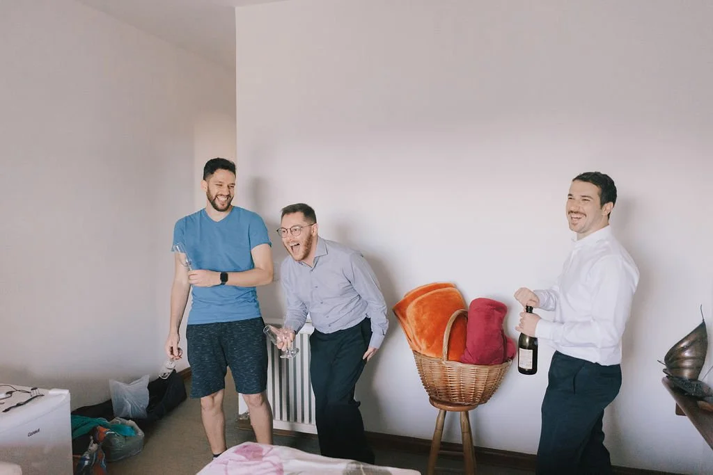 Três homens se divertindo em uma sala, um deles segura uma garrafa de bebida, outro segura um copo e o terceiro segura uma garrafa de vinho, com balões de festa ao fundo.