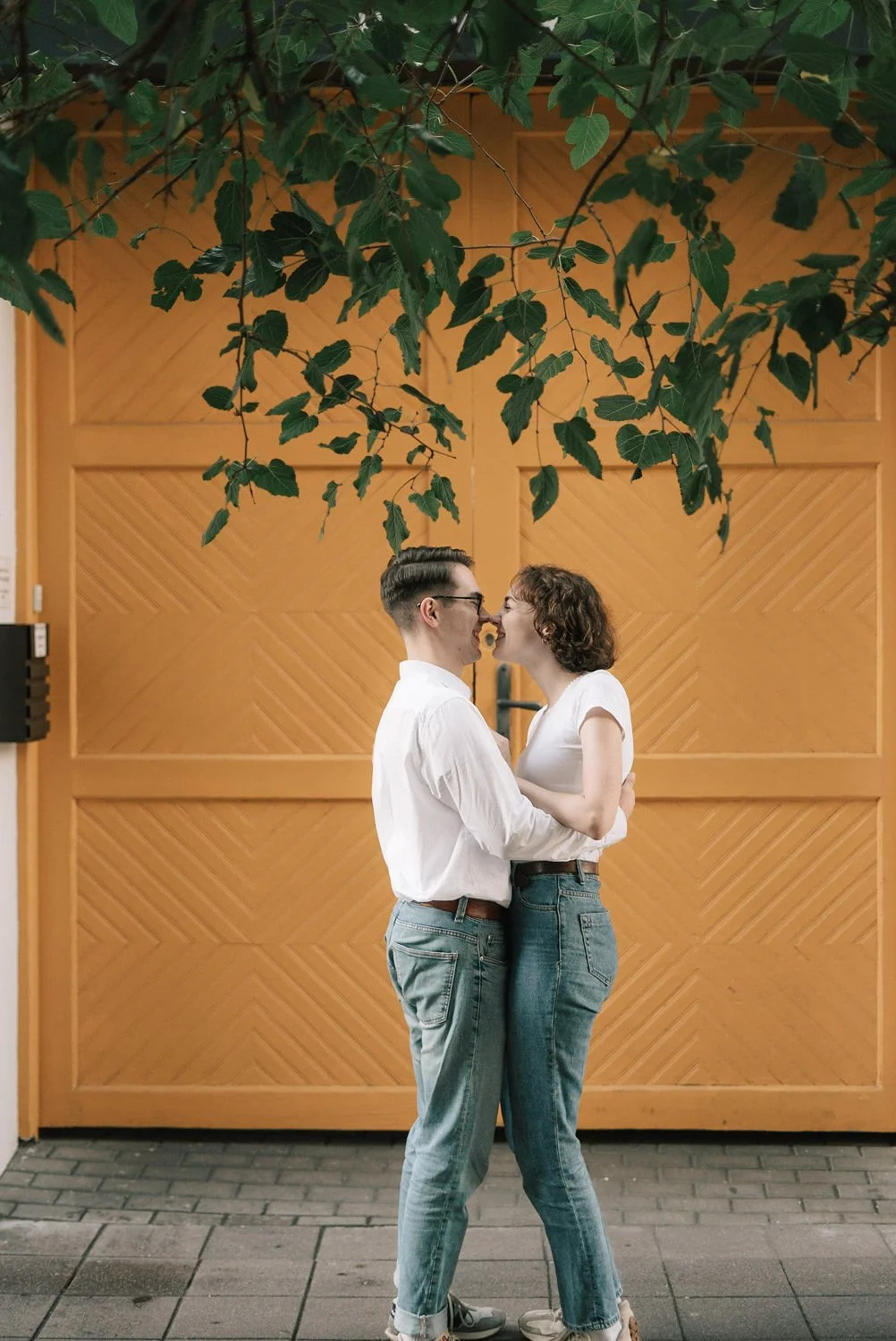 Um casal de mãos dadas brincando de fecha olhos, em frente a uma porta de madeira, sob árvores com folhas verdes.