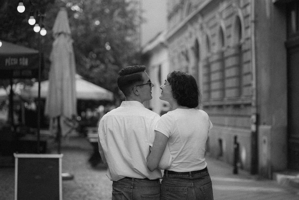 Homens e mulheres caminhando juntos na rua em um ambiente urbano ao entardecer, com prédios, iluminação de lâmpadas e guarda-sóis ao fundo.