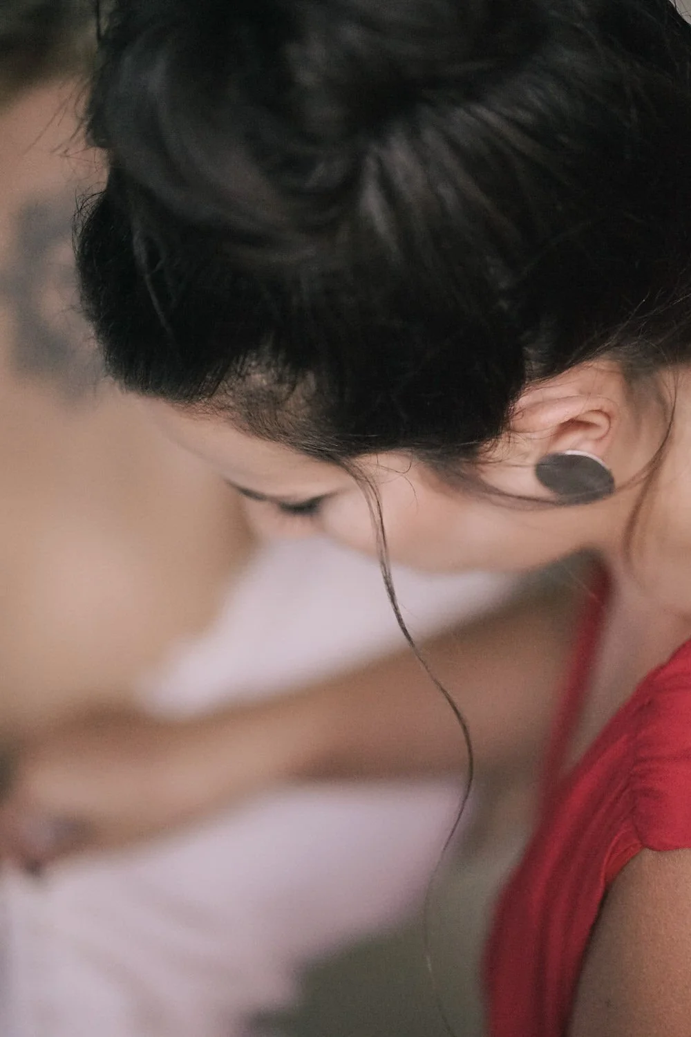 Close-up de duas pessoas com a cabeça abaixada, uma de cabelo escuro e outra de cabelo castanho, usando brincos grandes e roupas vermelhas.