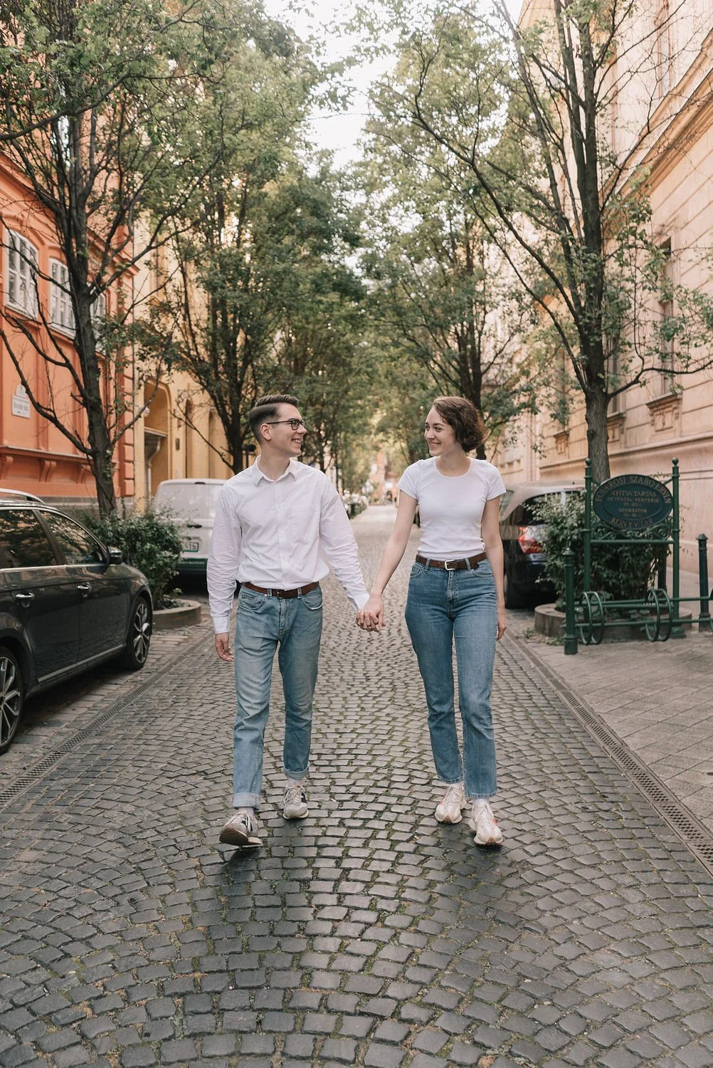 Casal caminhando de mãos dadas na rua pavimentada de paralelepípedo, cercada por árvores e edifícios antigos, em clima de descontração e alegria.