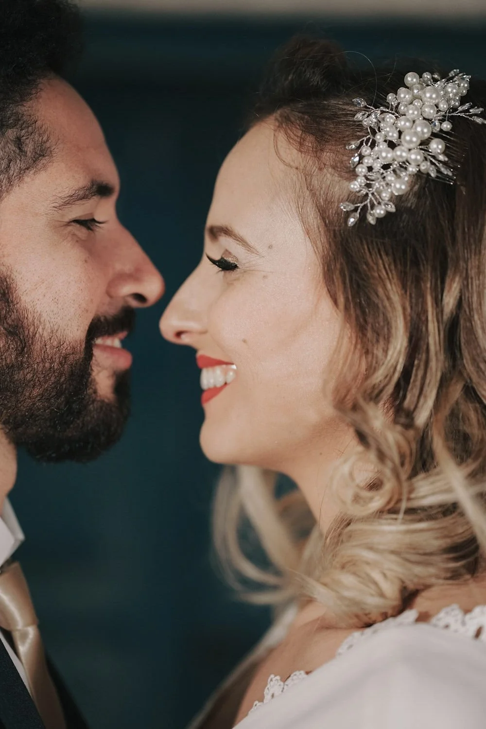 Casal em casamento, rosto bem próximo, sorrindo, com detalhes de maquiagem e acessórios de cabelo.