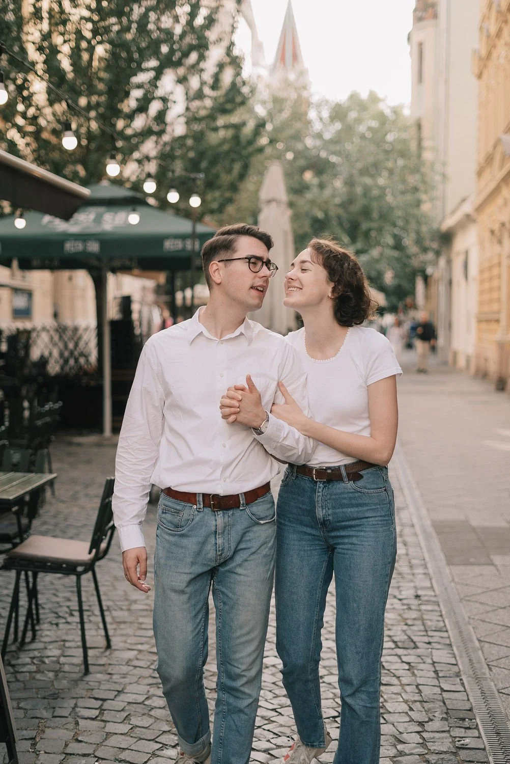 Um casal andando de mãos dadas em uma rua de paralelepípedos, sorrindo e olhando um para o outro, com árvores e prédios históricos ao fundo em um ambiente ao ar livre e ensolarado.