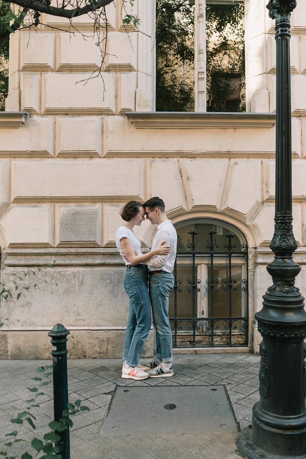 Um casal jovem, vestindo roupas casuais, está sorrindo e se tocando na rua, num ambiente urbano com uma parede de prédio antiga ao fundo.