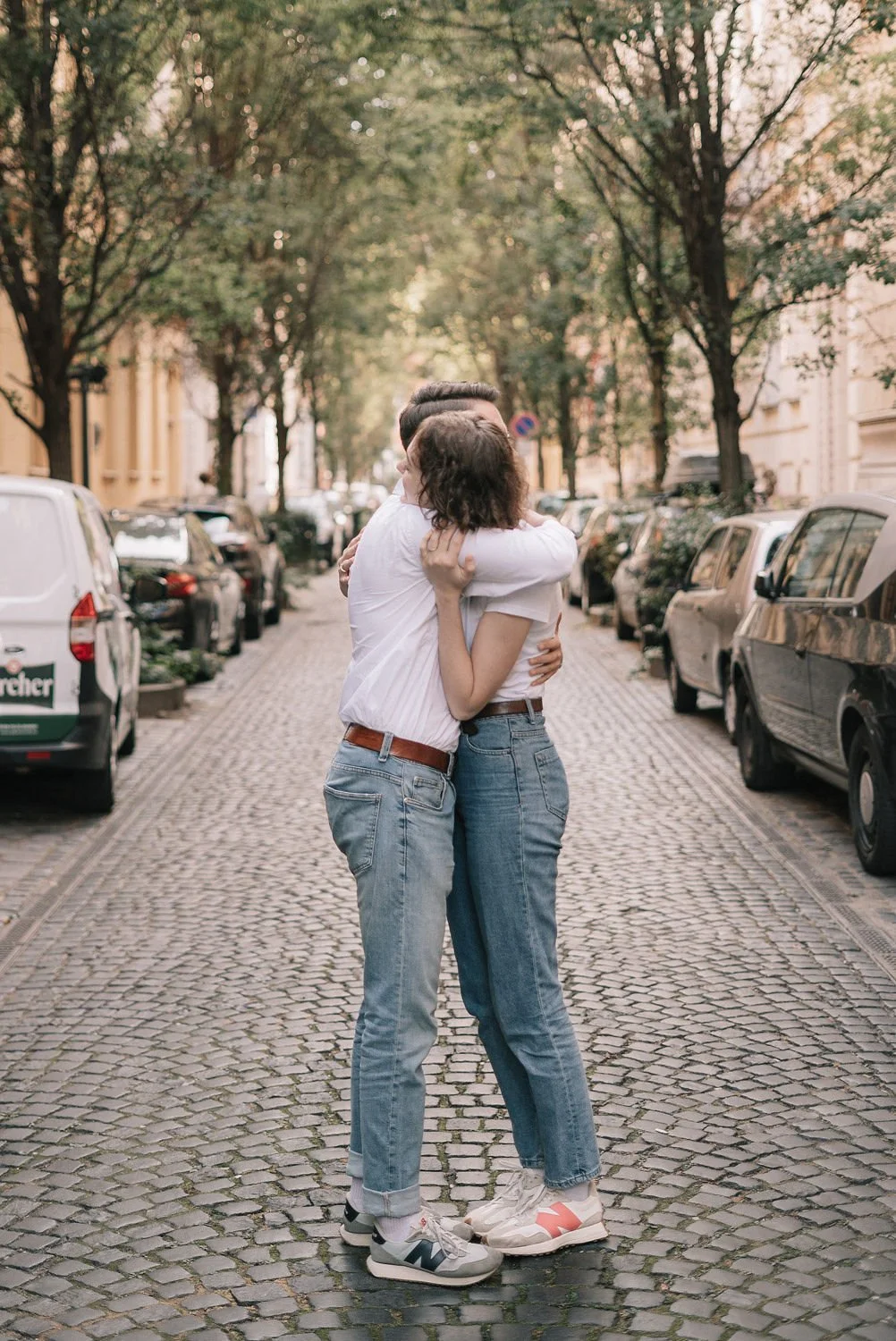 Casal se abraçando em rua de paralelepípedos, com árvores e carros estacionados ao redor.