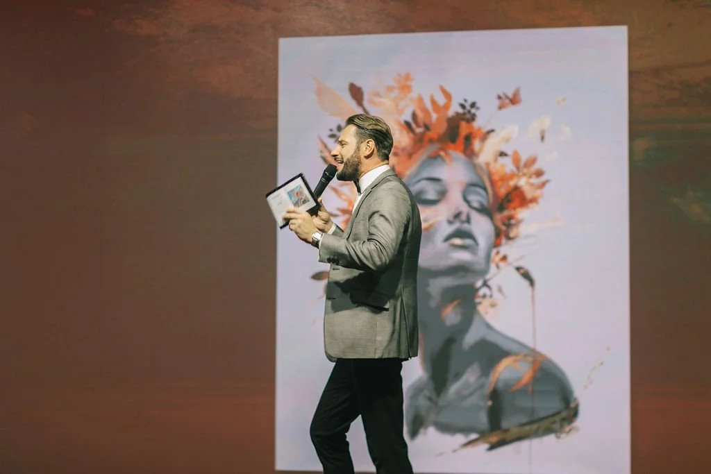 Homem de terno cinza apresentando com microfone e tablet, ao lado de uma arte de mulher com elementos florais na cabeça.