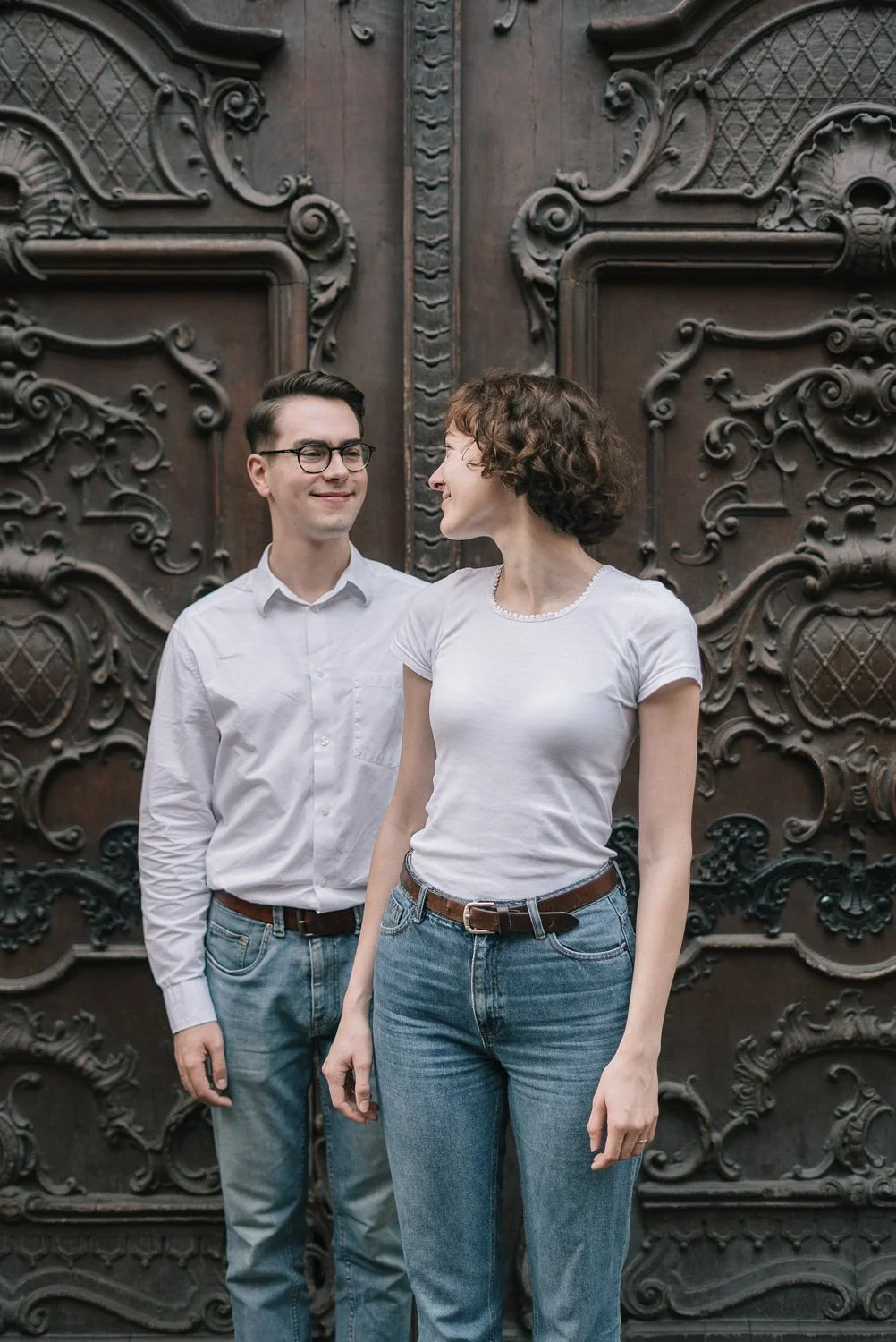 Um casal sorridente, de roupas casuais, de pé na frente de uma porta de madeira ornamentada, olhando um para o outro.